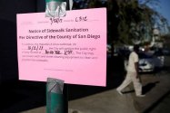 In this Sept. 28, 2017 file photo a man passes behind a sign warning of an upcoming street cleaning in San Diego. Health officials say a deadly California outbreak of Hepatitis A may take a year or more to abate.